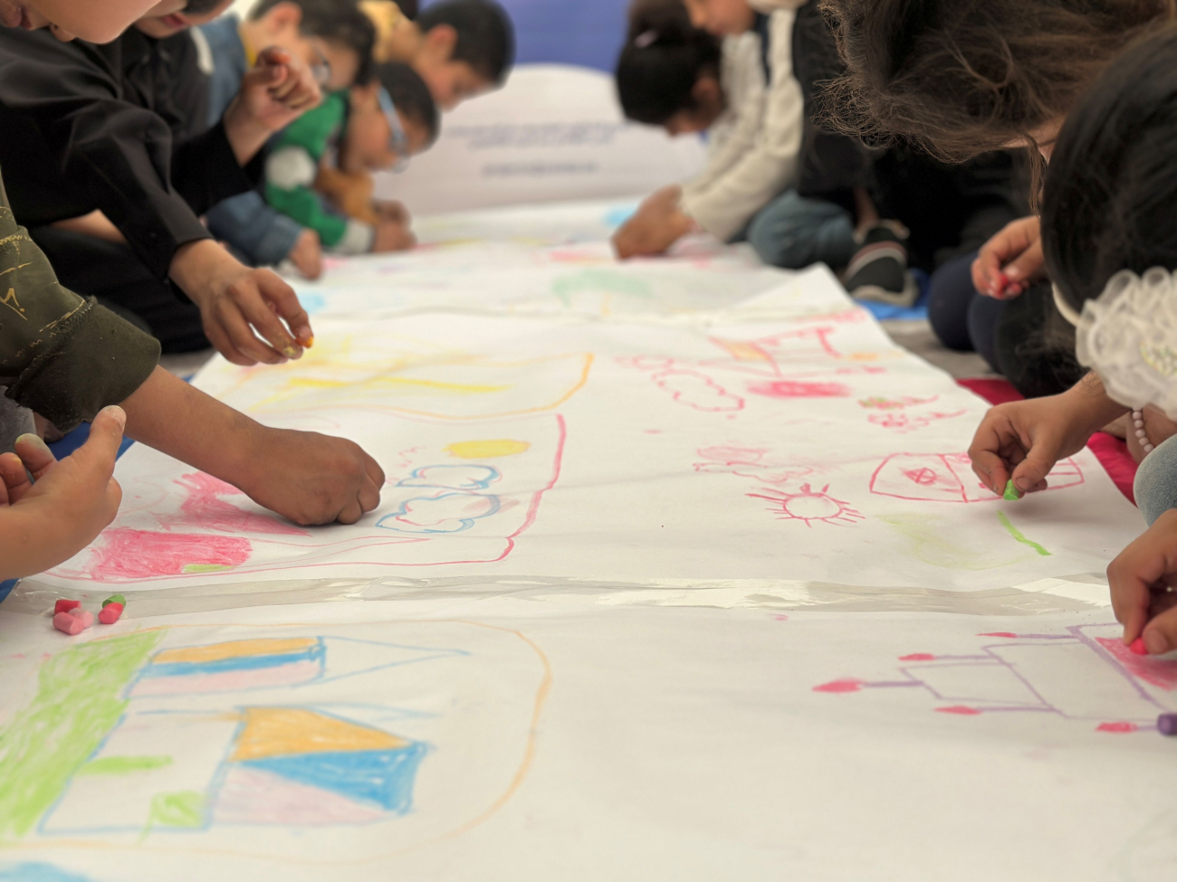 Kinder zeichnen mit bunten Stiften auf einer langen Papierrolle, die am Boden aufgelegt ist.